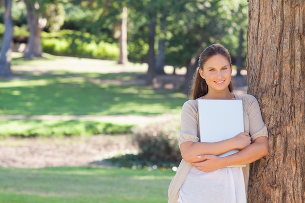 Smiling woman with a laptop leaning against a tree