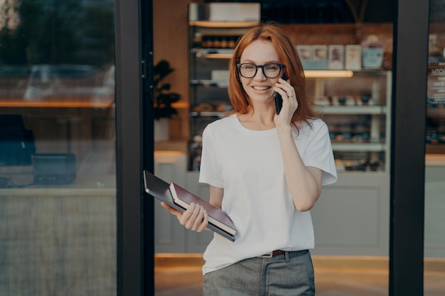 ラップトップとノートブックを手に笑顔の女性がコーヒーショップの近くで携帯電話で話を楽しんでいます