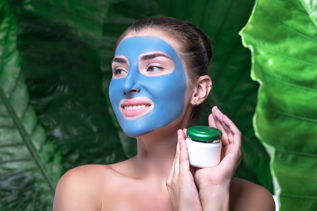 A smiling woman with a mask of blue clay on her face is holding a jar of cream.