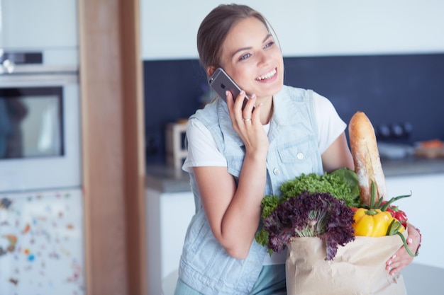 Smiling woman with mobile phone holding shopping bag in kitchen