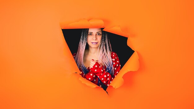 Photo smiling woman with senegalese braids in a hole in the wall a stylish girl in a bright red dress looks at the camera through a hole in the orange wall in the studio