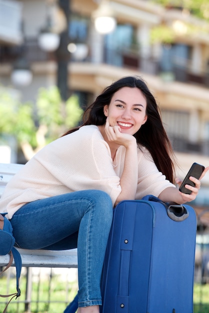 smiling woman with suitcase and cellphone sitting on park bench