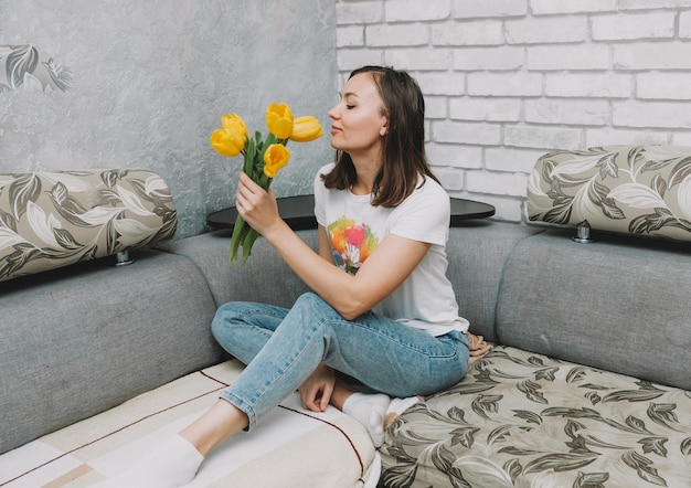 Smiling woman with yellow tulips is sitting on the sofa at home