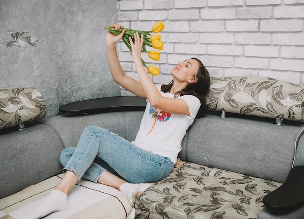 Smiling woman with yellow tulips is sitting on the sofa at home