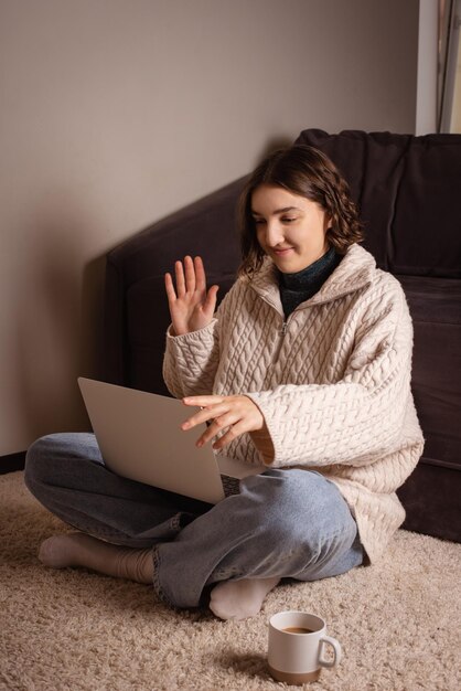 Photo smiling woman work at home on laptop on floor and wave hand to greet coworkers in online meeting