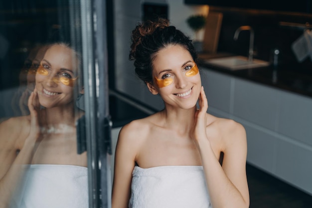 Smiling woman wrapped in towel using under eye patches after shower at home Skincare treatment