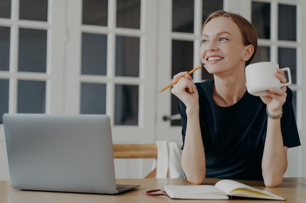 Smiling woman write notes in notebook sitting at desk thinking planning day visualizing dreams