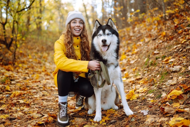 黄色いコートを着た笑顔の女性が、晴れた天気の秋の森でかわいいペットのハスキーと一緒に歩きます