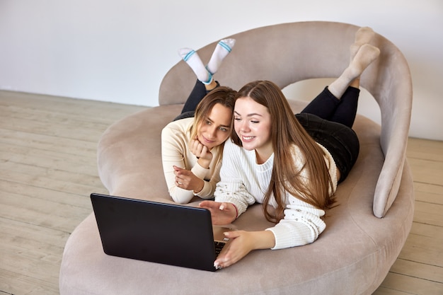 Smiling women look at laptop while lie on settee in lounge