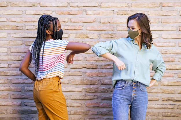 Photo smiling women wearing mask doing elbow bump standing against wall