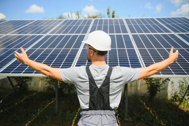 Smiling worker with solar station raising his hands showing thumbs up on a background of photovoltaic panels near the house Man in orange uniform Science solar energy Renewable energy