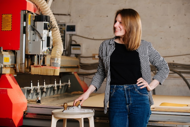 Smiling worker woman in semi official clothes standing next to
a big red piece of heavy machinery. she placed her hand on a small
plane on a stool.