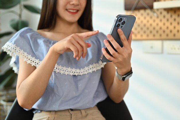 笑顔の若い成人アジア人女性が、スマートフォンでオンラインでクライアントとチャットし、職場でラップトップを使用しています