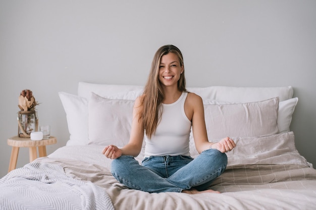 Smiling young adult Italian woman in white tshirt sitting on bed in yoga meditation pose looks