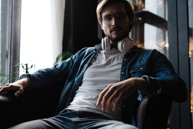 smiling young adult man wearing wireless headphones listening mobile music on smartphone, happy guy using online player podcast audio book in app on cell phone relaxing at home