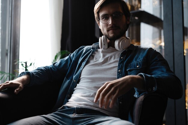 Smiling young adult man wearing wireless headphones listening mobile music on smartphone, happy guy using online player podcast audio book in app on cell phone relaxing at home