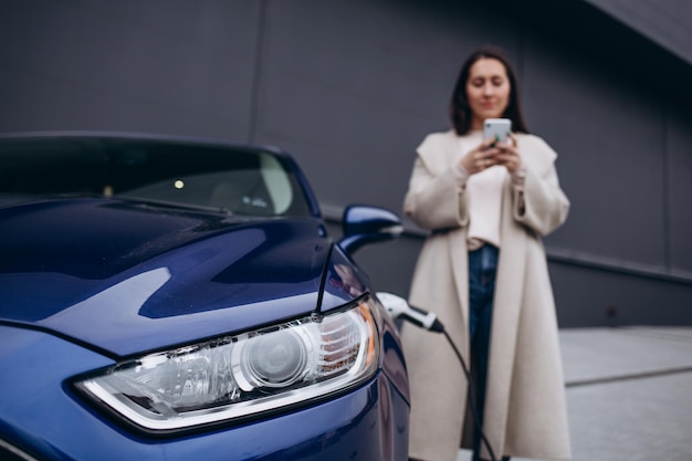 Smiling young adult woman standing on city parking near\
electric car charging automobile battery from small city station\
drinking coffee and talking on smartphone