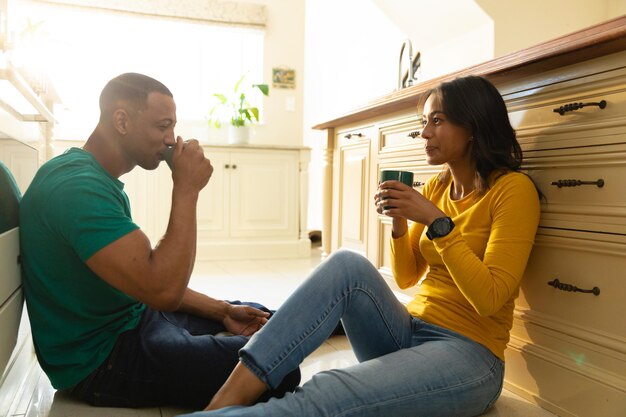 Photo smiling young african american couple drinking coffee while sitting on kitchen floor at home. people, love and togetherness concept, unaltered.