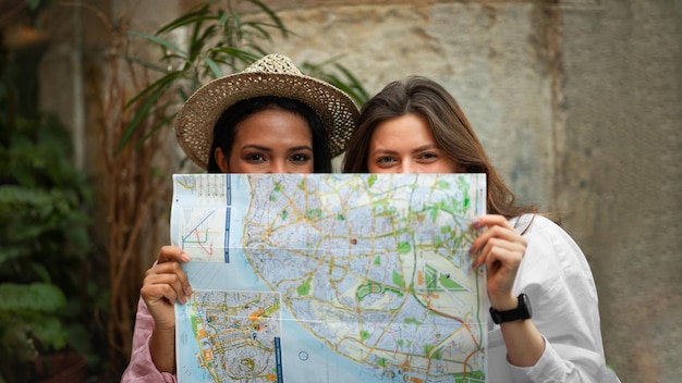 Smiling young african american and european women in hat looking for route peeking out from behind map