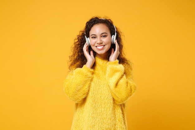 Smiling young african american girl in fur sweater posing isolated on yellow orange background in studio. People sincere emotions lifestyle concept. Mock up copy space. Listen music with headphones.