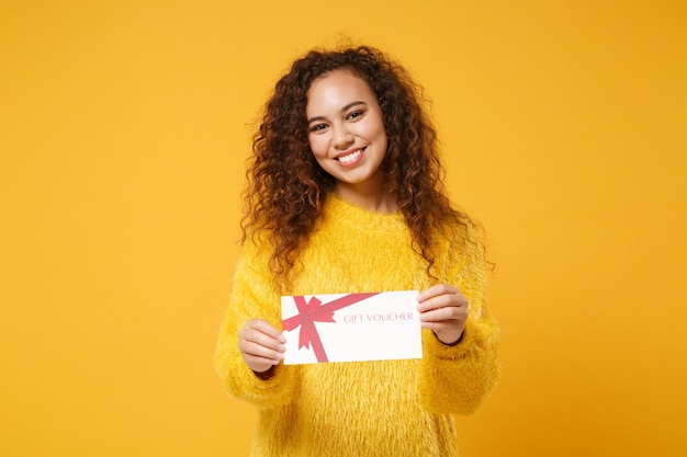 Smiling young african american girl in fur sweater posing isolated on yellow orange background studio portrait. People lifestyle concept. Mock up copy space. Hold gift certificate, keeping mouth open.