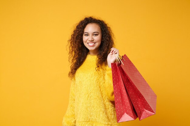 Smiling young african american girl in fur sweater posing isolated on yellow orange wall background. people lifestyle concept. mock up copy space. holding package bag with purchases after shopping.