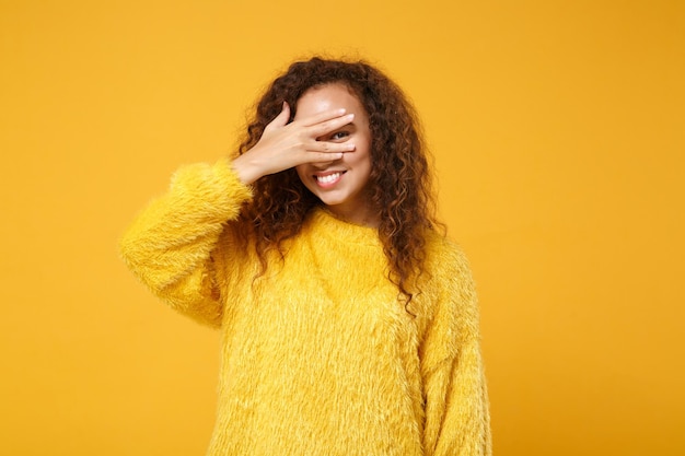 Smiling young african american girl in fur sweater posing isolated on yellow orange wall background, studio portrait. People lifestyle concept. Mock up copy space. Covering face with hand, peeping.