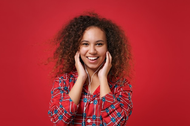 Smiling young african american girl in pajamas homewear posing while resting at home isolated on red background. relax good mood lifestyle concept. mock up copy space. listen music with earphones