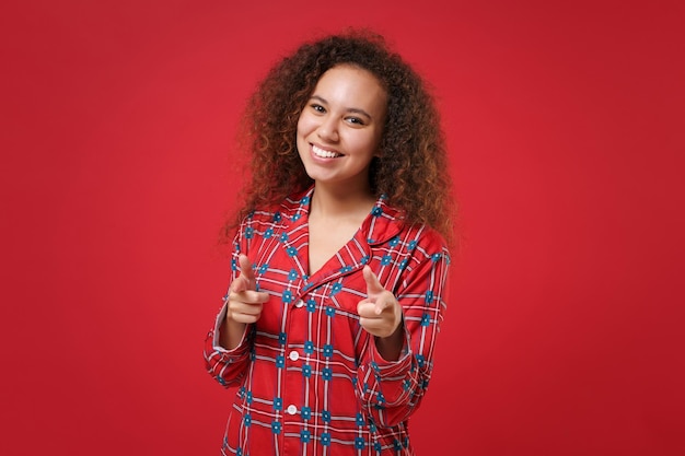 Улыбающаяся молодая афро-американская девушка в домашней одежде пижамы позирует во время отдыха дома на красном фоне. Расслабьтесь, концепция образа жизни в хорошем настроении. Скопируйте пространство для копирования. Наведите указательные пальцы на камеру.