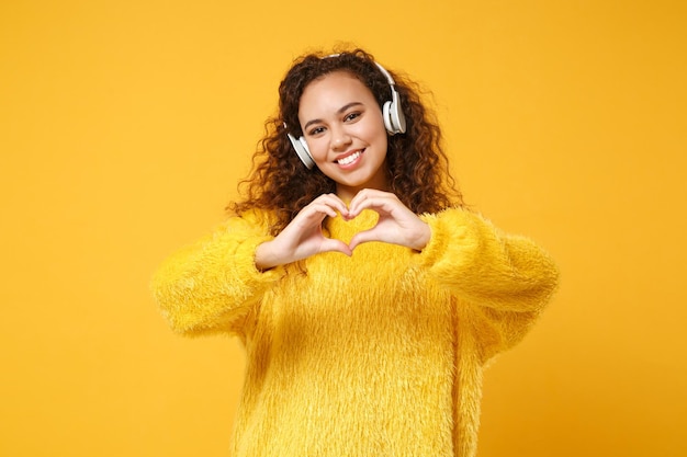 Улыбающаяся молодая афроамериканская девушка в свитере позирует изолированной на желто-оранжевом фоне. Концепция образа жизни людей. Скопируйте пространство для копирования. Слушайте музыку в наушниках, показывая руками форму сердца.
