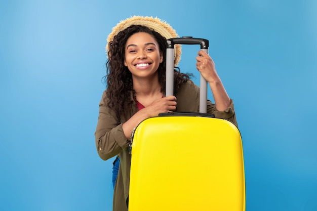 Smiling young african american lady sitting behind bright yellow suitcase going on summer vacation