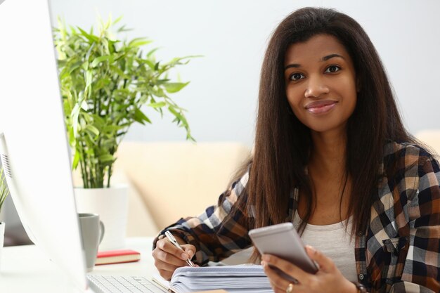 Smiling young african american woman writing in notebook studying on mobile phone online