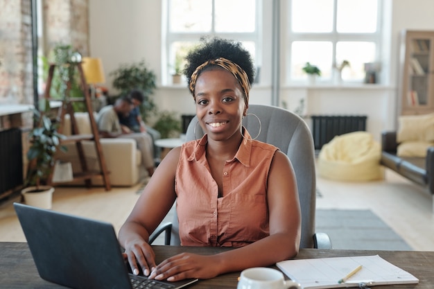 Smiling young African female freelancer looking at you during work in front of laptop while sitting in armchair by table in front of camera