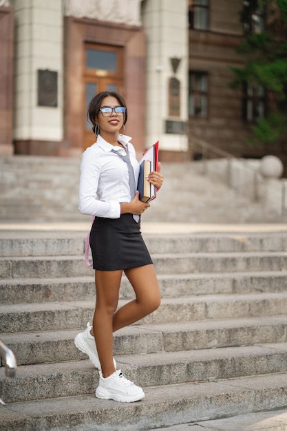 Smiling young African girl in eyeglasses with books on the stairs near the school or college