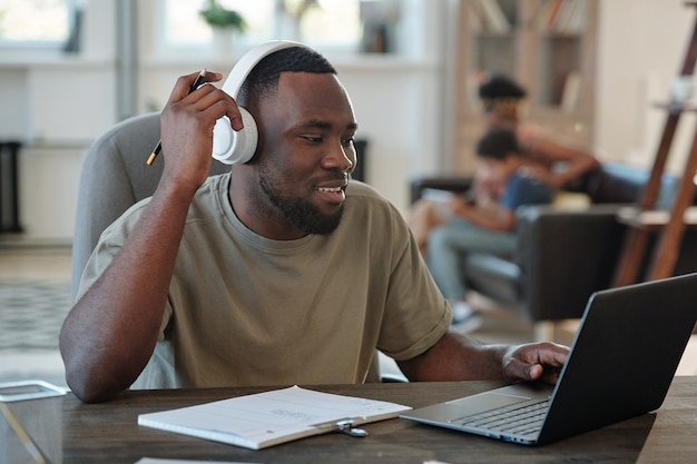 Smiling young African man in headphones sitting by table in front of laptop and scrolling through online data against his family