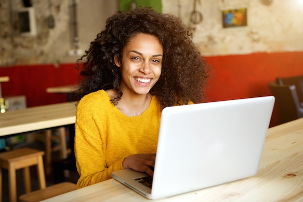 Giovane donna africana sorridente che si siede in un caffè con il computer portatile