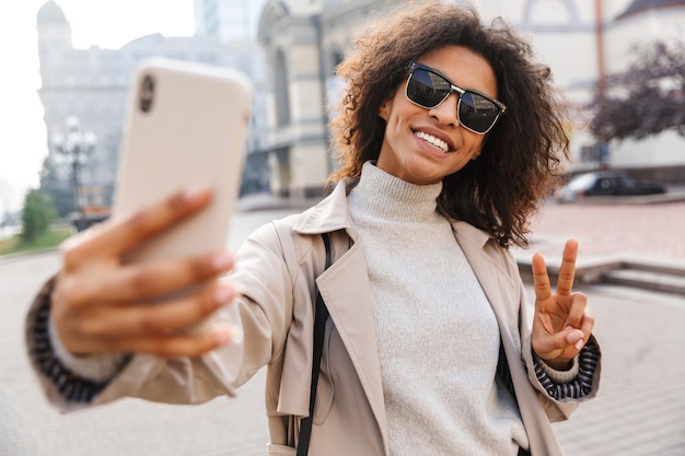 Sorridente giovane donna africana che indossa un cappotto autunnale che cammina all'aperto, facendo un selfie