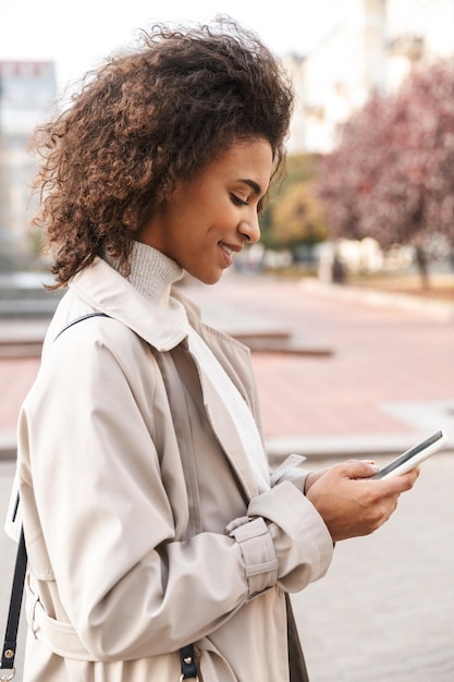 Smiling young african woman wearing autumn coat walking outdoors, using mobile phone