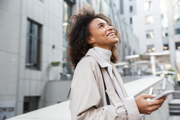 Photo smiling young african woman wearing autumn coat walking outdoors, using mobile phone