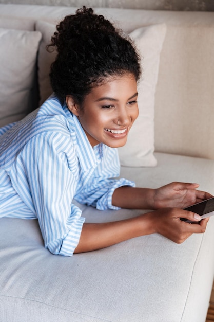横たわっている間携帯電話を使用して、自宅のソファでリラックスしたシャツを着て笑顔の若いアフリカの女性