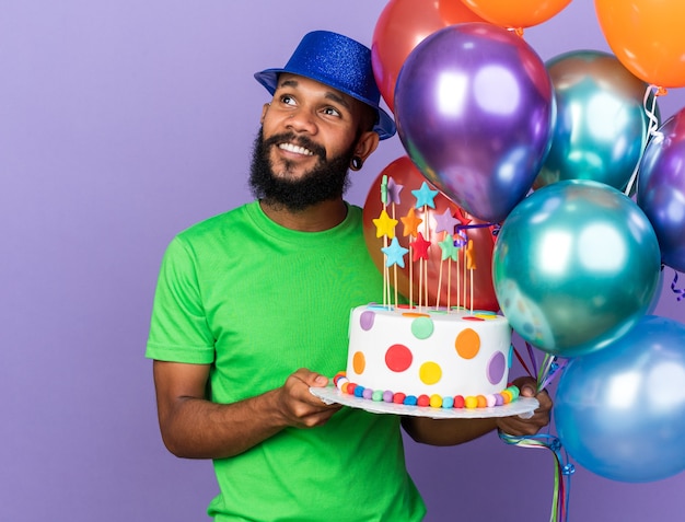 Smiling young afro-american guy wearing party hat holding balloons with cake isolated on blue wall with copy space