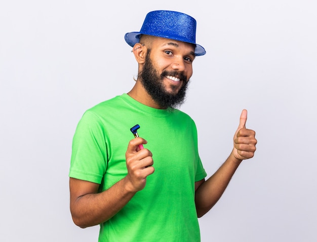 Smiling young afro-american guy wearing party hat showing thumb up holding party whistle isolated on white wall