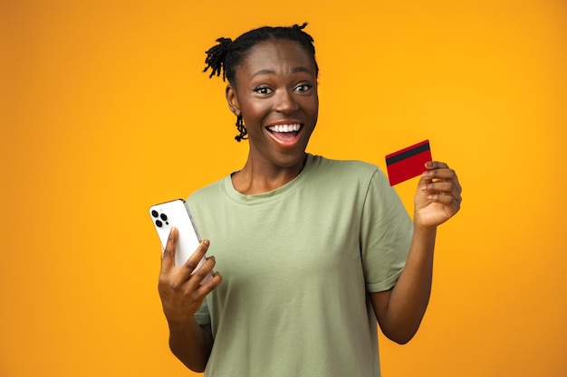 Smiling young afro girl holds smart phone and credit card in yellow studio