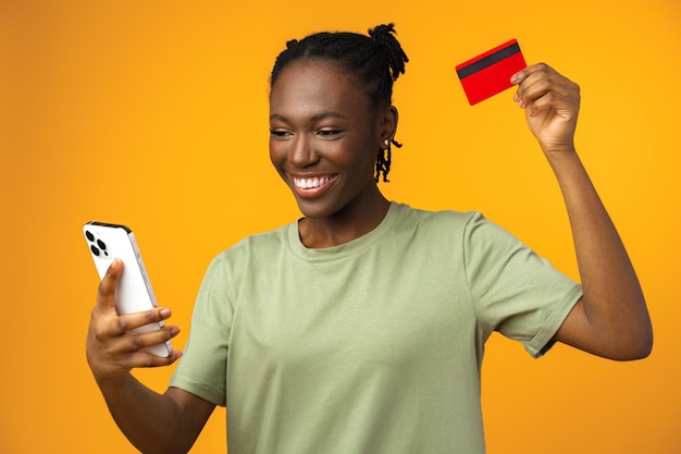 Smiling young afro girl holds smart phone and credit card in yellow studio