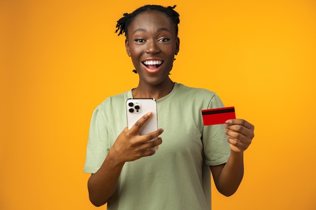 La giovane ragazza afro sorridente tiene lo smartphone e la carta di credito in studio giallo