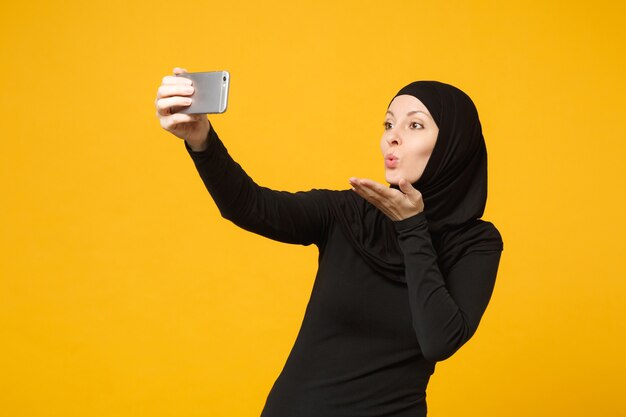 Smiling young arabian muslim woman in hijab black clothes doing selfie shot on mobile phone isolated on yellow wall  portrait. people religious lifestyle concept. .