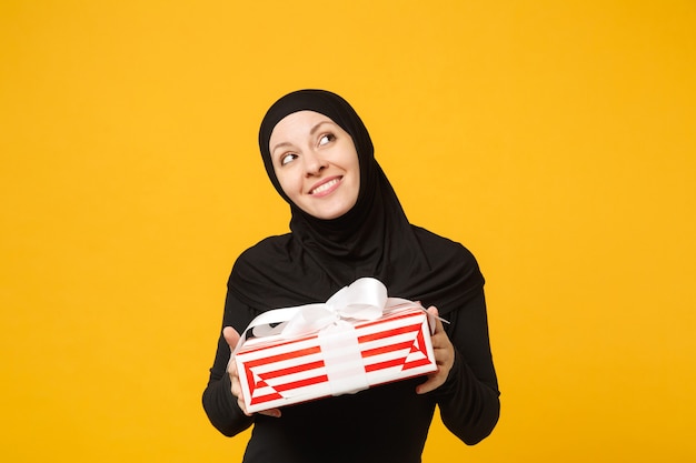 Smiling young arabian muslim woman in hijab black clothes hold in hand present box with gift isolated on yellow wall portrait. people religious lifestyle concept