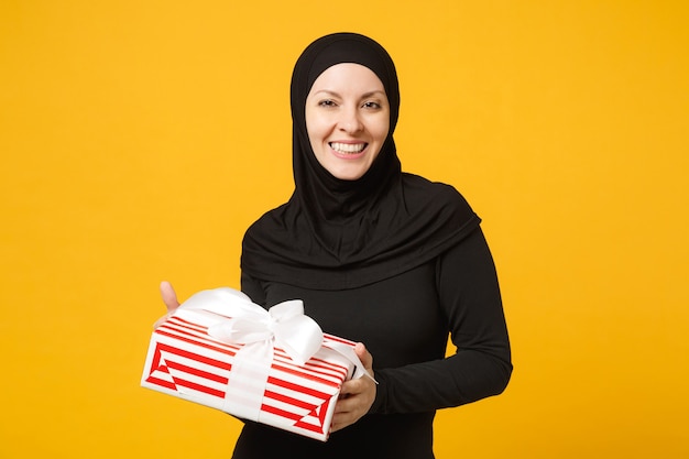 Smiling young arabian muslim woman in hijab black clothes hold in hand present box with gift isolated on yellow wall  portrait. People religious lifestyle concept. .