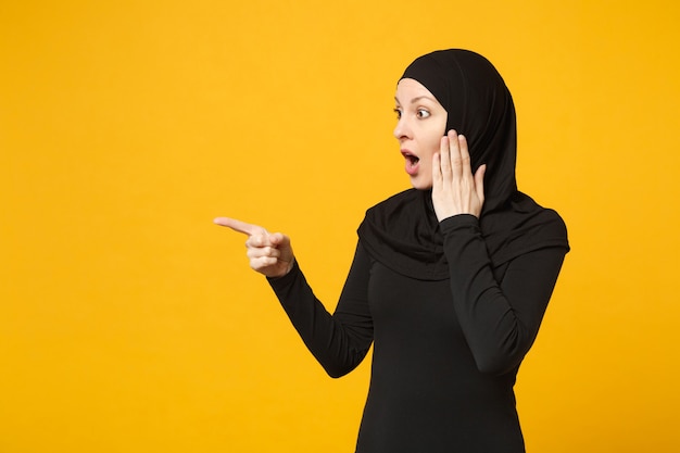 노란색 벽 초상화에 고립 된 손 손가락으로 복사 공간을 가리키는 hijab 검은 옷에 젊은 아라비아 무슬림 여성 미소. 사람들이 종교적인 라이프 스타일 개념.