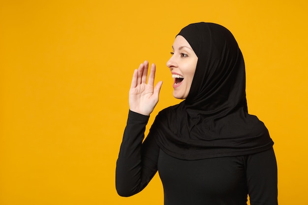 Smiling young arabian muslim woman in hijab black clothes whispering secret behind her hand isolated on yellow wall, portrait. People religious lifestyle concept. 